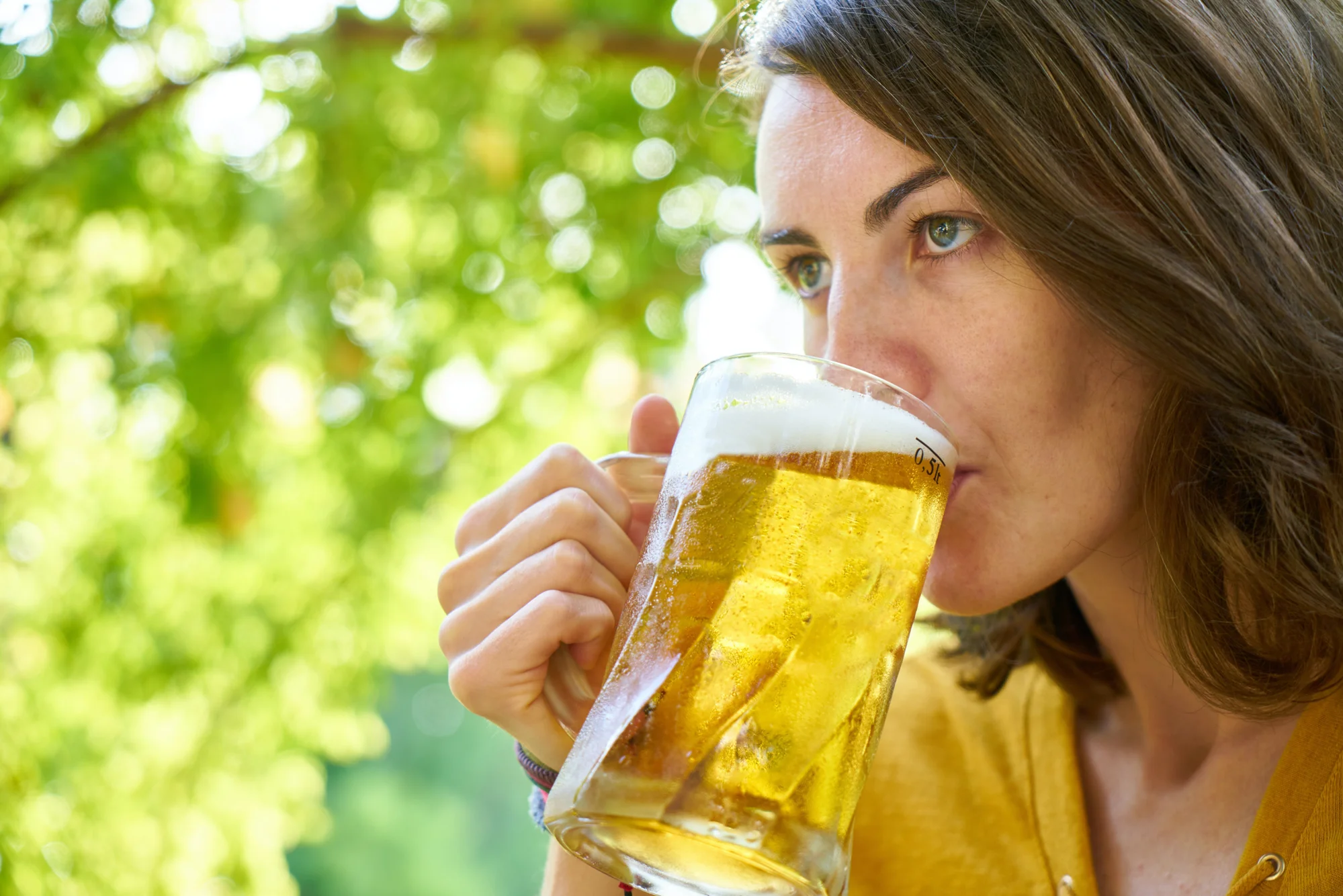 Mujer joven bebe cerveza de una jarra de medio litro en un entorno natural con vegetación de fondo.
