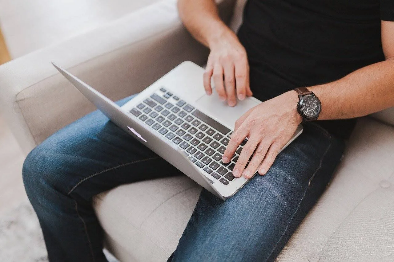 Una persona con remera negra y jeans escribe en una laptop apoyada sobre sus piernas mientras está sentada en un sofá.