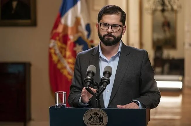 Hombre con lentes y traje gris habla desde un podio con el escudo de Chile, ante micrófonos, con la bandera chilena al fondo.
