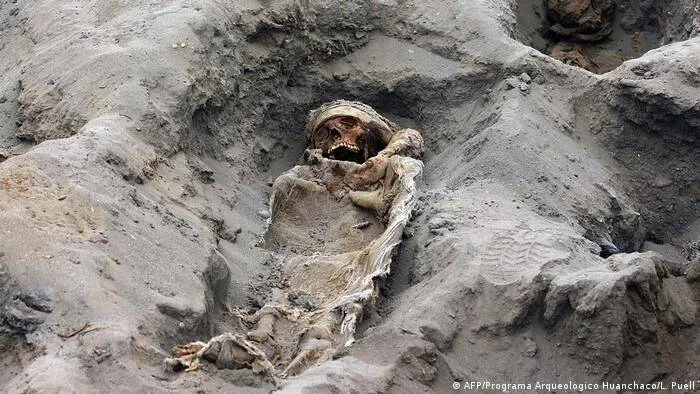 Esqueleto momificado parcialmente enterrado en sedimento seco, hallado en excavación arqueológica en Huanchaco, Perú.