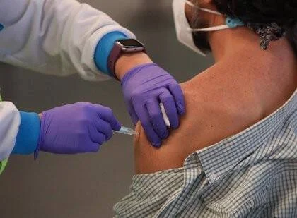 Un profesional de salud con guantes morados aplica una inyección en el brazo de una persona con camisa a cuadros.