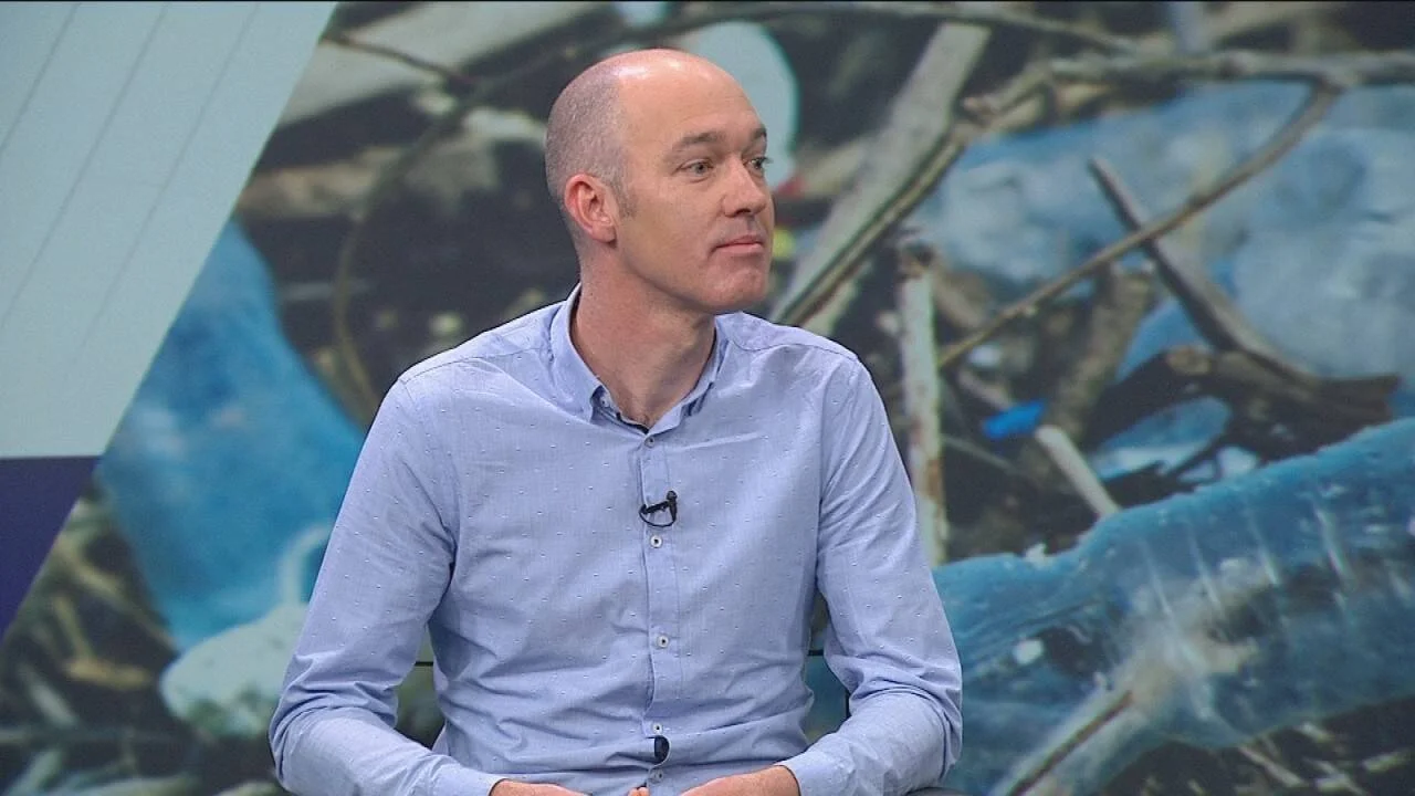 Hombre con camisa celeste y micrófono de solapa en estudio de televisión, con imagen de residuos plásticos de fondo.