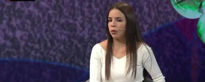 Mujer de cabello largo y remera blanca habla con micrófono de solapa en un estudio de televisión de fondo violeta.