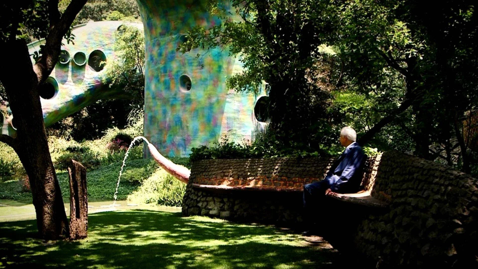 Hombre mayor sentado en un banco de piedra observa una estructura arquitectónica iridiscente con formas orgánicas y fuente de agua en un jardín arbolado.