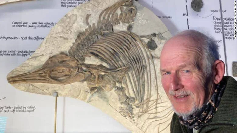 Un hombre mayor posa sonriente junto a un gran fósil de ictiosaurio exhibido en una pared con anotaciones científicas.