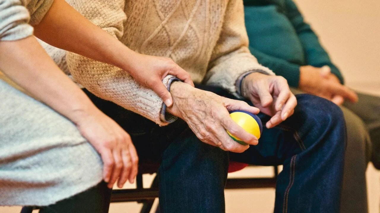 Una mano joven sostiene el brazo de una persona mayor que sujeta una pelota de colores, en un contexto de cuidado o rehabilitación.