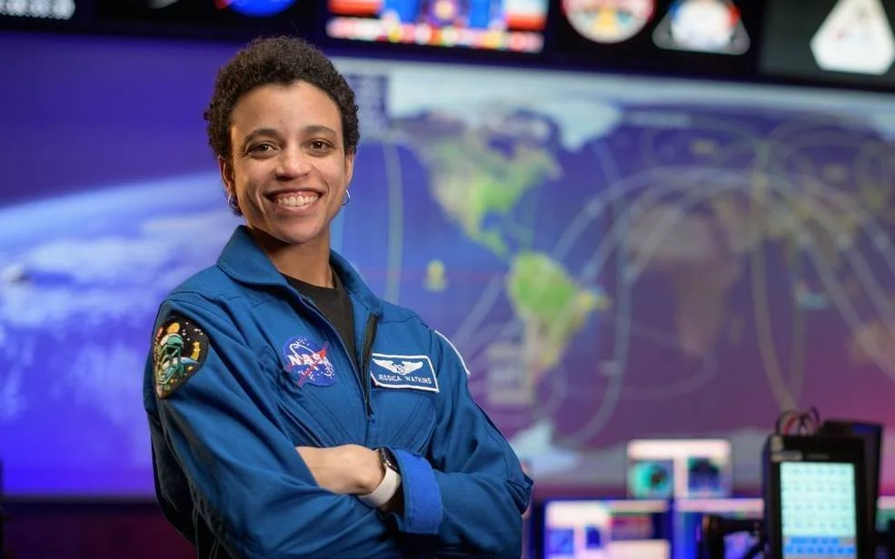 Astronauta con traje azul de la NASA, brazos cruzados, sonriendo frente a pantallas con mapa del mundo en centro de control espacial.