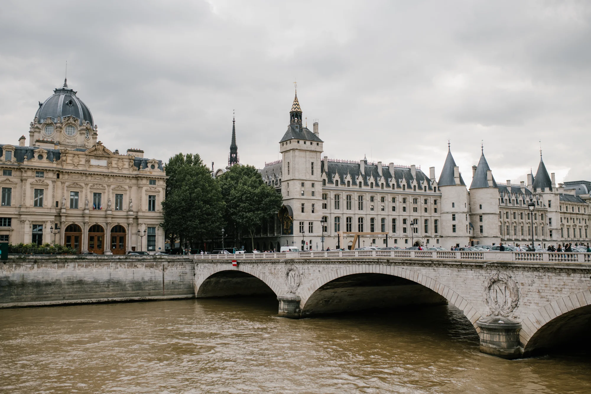 Puente de piedra sobre el río Sena con la Conciergerie y edificios neoclásicos de París al fondo bajo cielo nublado.