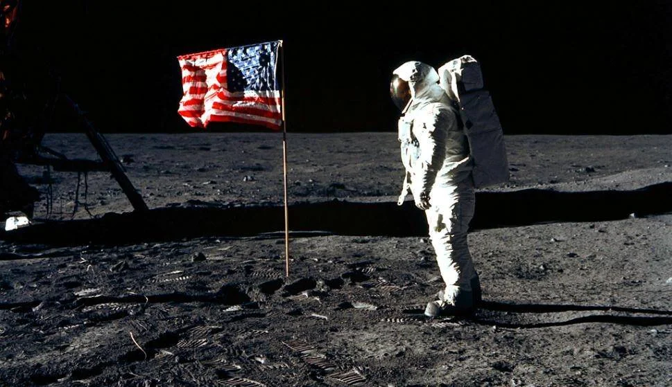 Astronauta con traje espacial de pie junto a la bandera de Estados Unidos plantada en la superficie lunar.