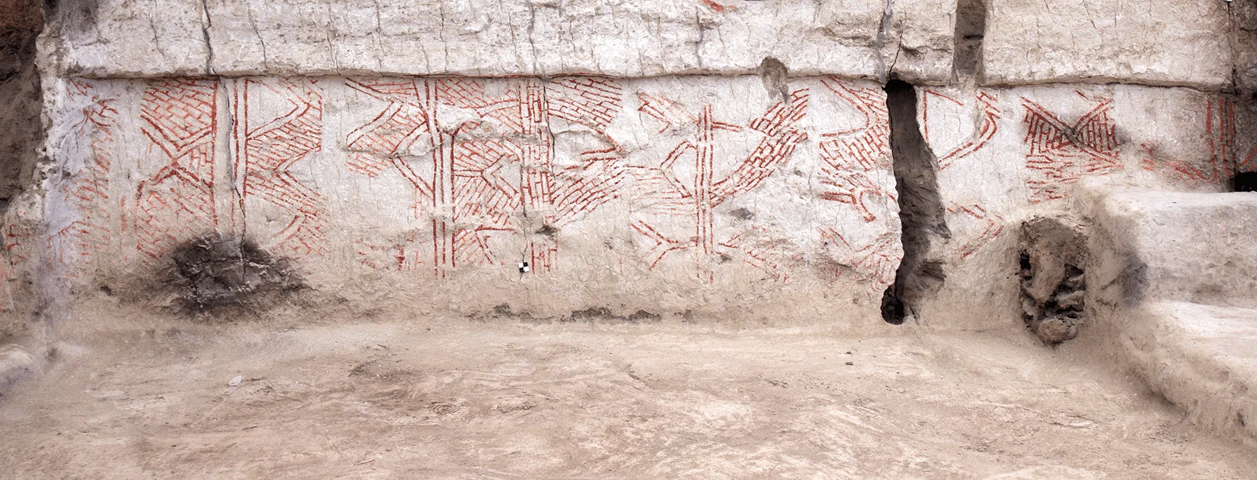 Muro de adobe con pinturas murales en pigmento rojo que representan figuras geométricas y animales en un sitio arqueológico.