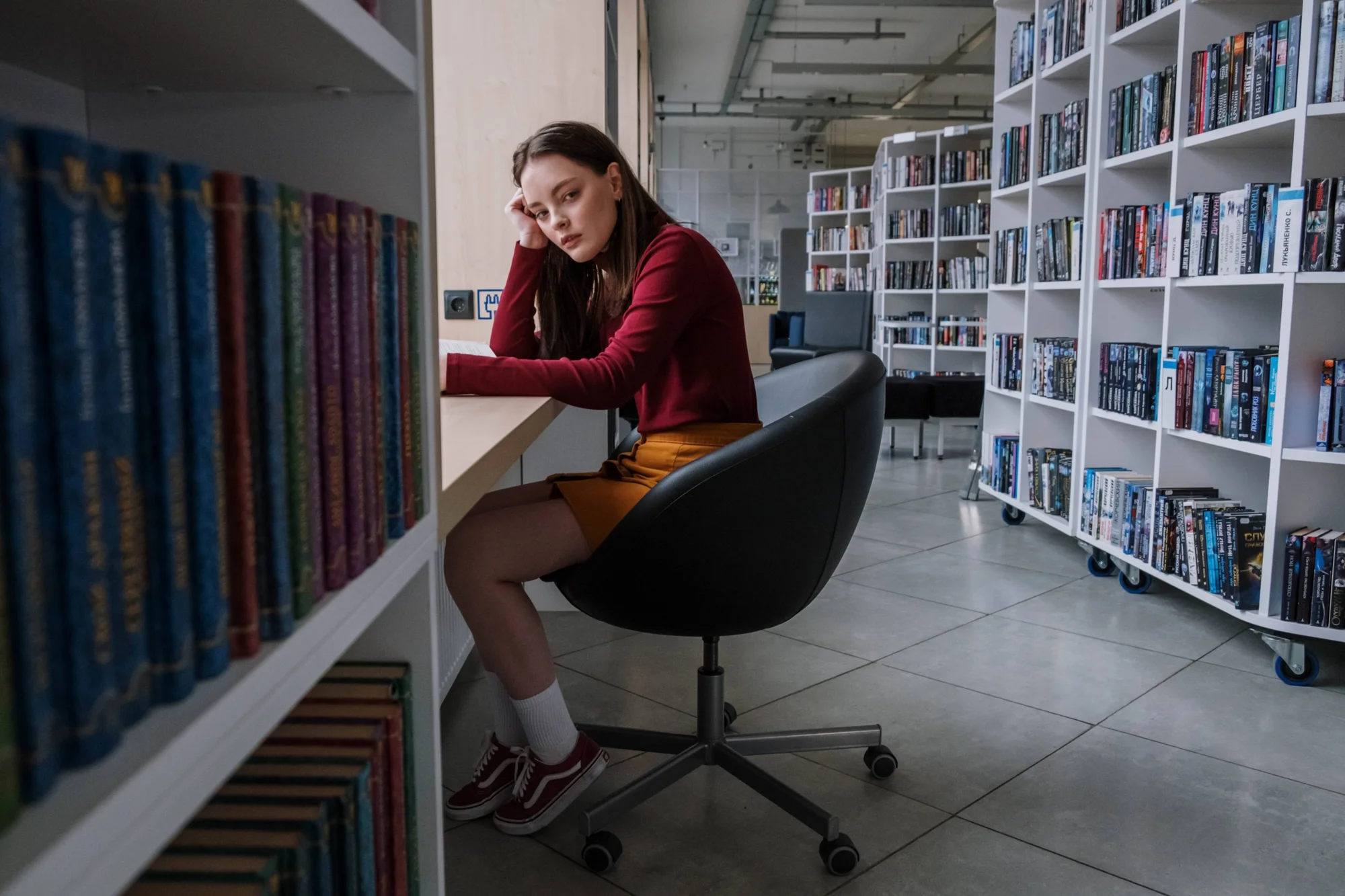 Joven con expresión pensativa apoya la cabeza en su mano mientras está sentada en una silla de escritorio en una biblioteca rodeada de estantes con libros.