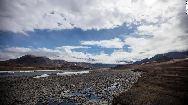 Lecho de río pedregoso con fragmentos de hielo en un valle ártico rodeado de montañas bajo cielo nublado.