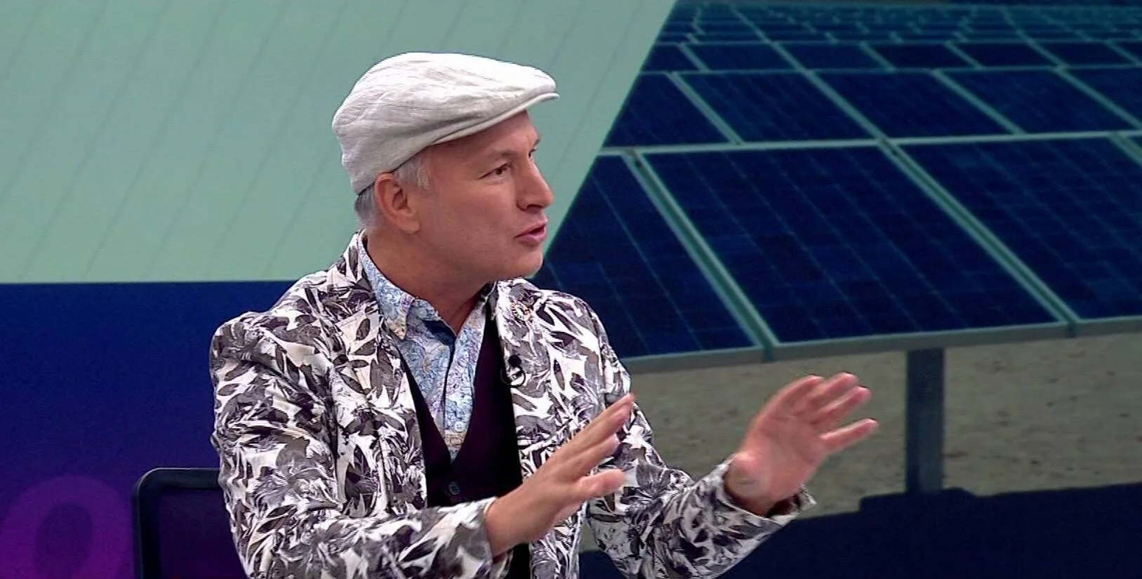 Hombre con boina blanca y saco estampado gesticula mientras habla en un estudio televisivo con paneles solares de fondo.