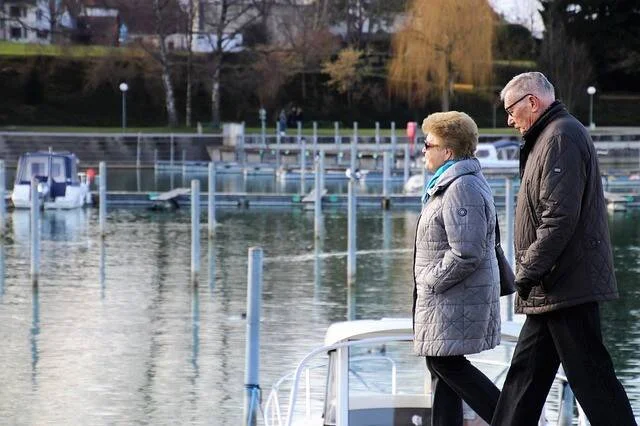 Dos personas mayores caminan junto a un muelle con embarcaciones y agua al fondo en un día soleado.