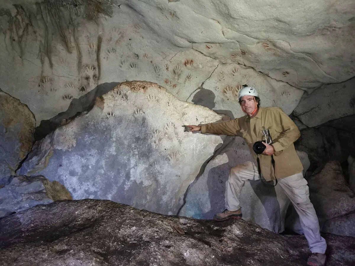 Un hombre con casco y cámara señala decenas de impresiones de manos prehistóricas en la pared de una cueva.