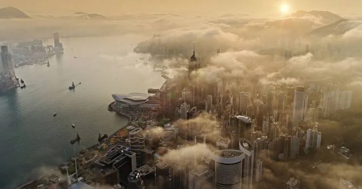 Panorama aéreo de una densa ciudad costera con rascacielos emergiendo entre nubes bajo una luz dorada al amanecer.