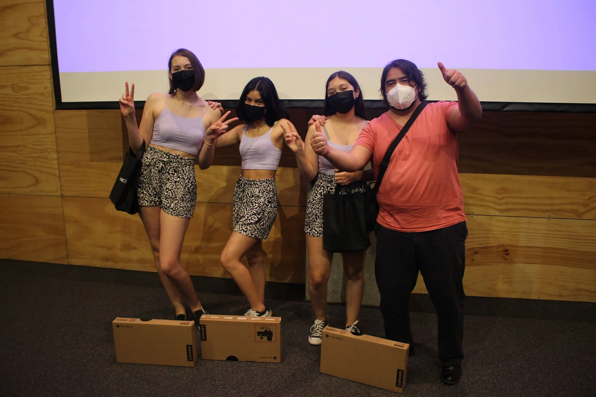 Cuatro jóvenes con mascarilla posan celebrando junto a cajas de laptops Lenovo en un auditorio.