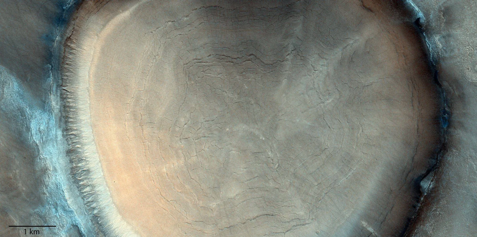 Vista aérea de un cráter en Marte con capas sedimentarias concéntricas y grietas en su suelo, escala de 1 km.