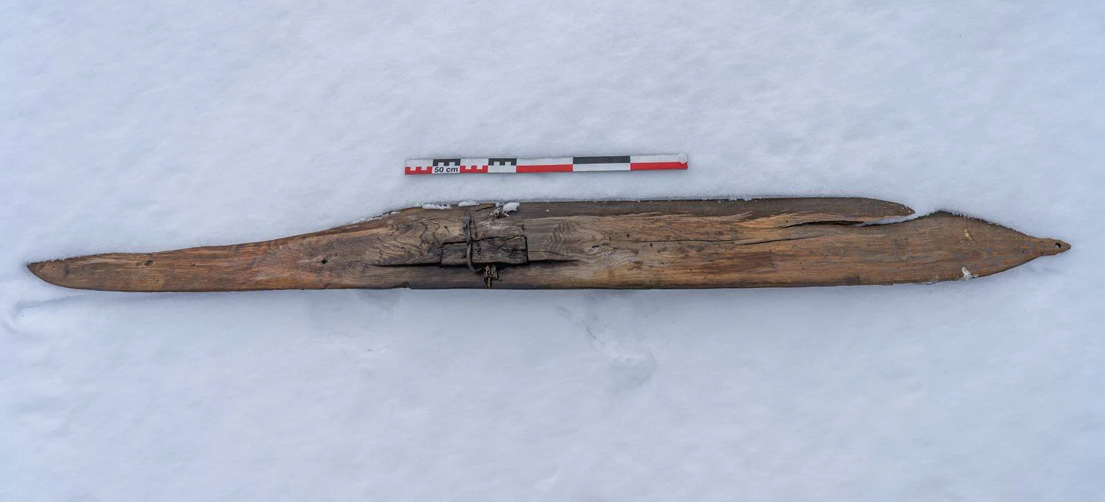 Esquí prehistórico de madera con ligadura central conservado sobre nieve, junto a escala métrica de 50 cm.