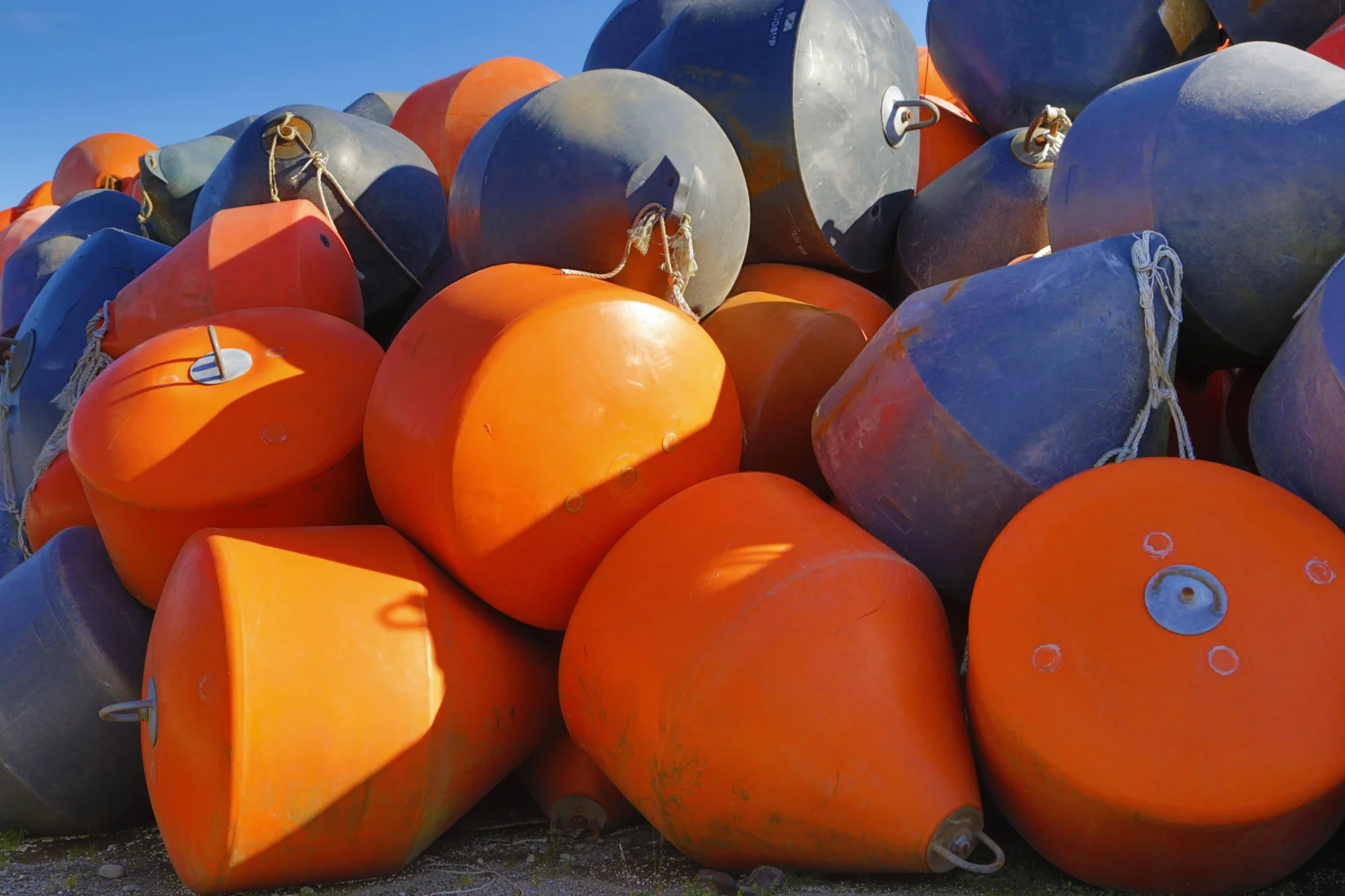 Gran cantidad de boyas marinas de colores naranja y gris apiladas en tierra bajo un cielo azul despejado.