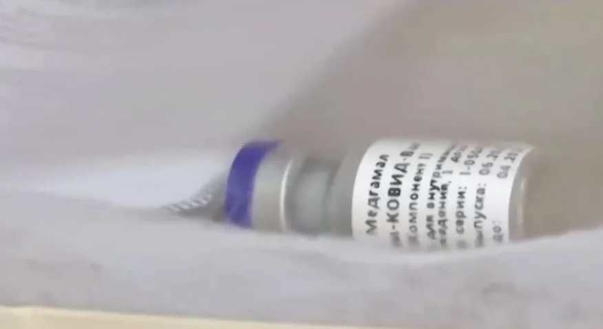Vial de vacuna con etiqueta en cirílico que incluye la palabra КОВИД (COVID), apoyado sobre una superficie blanca.