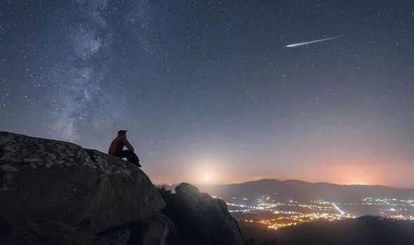 Una persona sentada en lo alto de una roca contempla un meteoro cruzando el cielo nocturno estrellado sobre un valle iluminado.