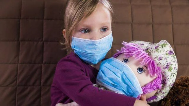 Niña pequeña con mascarilla quirúrgica abraza una muñeca de trapo que también lleva mascarilla.
