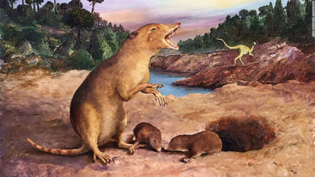 Reconstrucción artística de un mamífero prehistórico erguido con la boca abierta junto a dos crías, con dinosaurios al fondo.