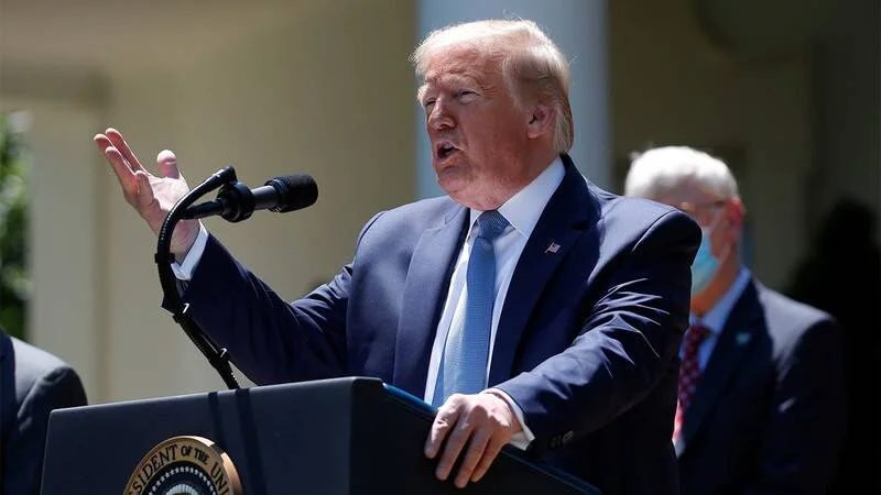 Hombre con traje azul gesticula mientras habla en un atril con el sello presidencial de Estados Unidos en un jardín.