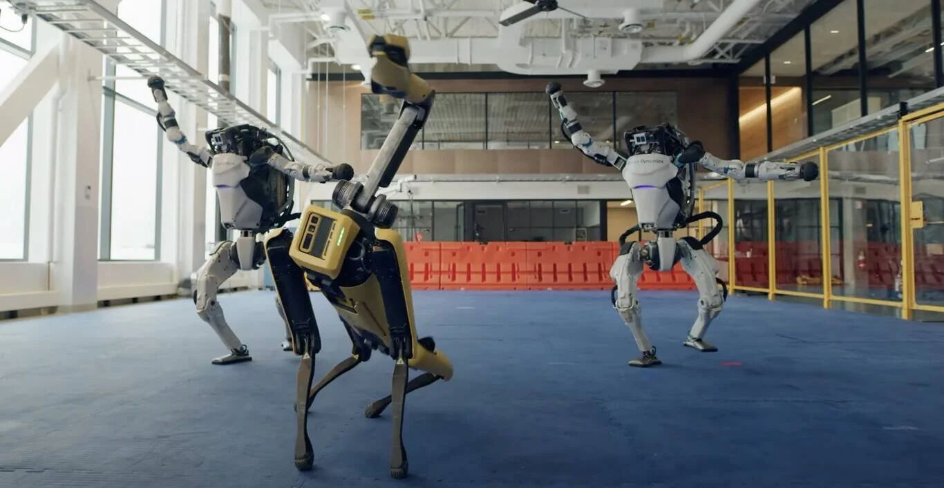 Dos robots humanoides y un robot cuadrúpedo amarillo realizan movimientos de baile sincronizados en un amplio espacio interior.