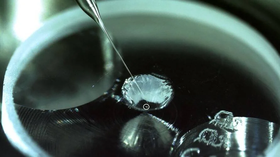 Una aguja delgada vierte una gota de líquido transparente sobre una superficie metálica reflectante en primer plano.