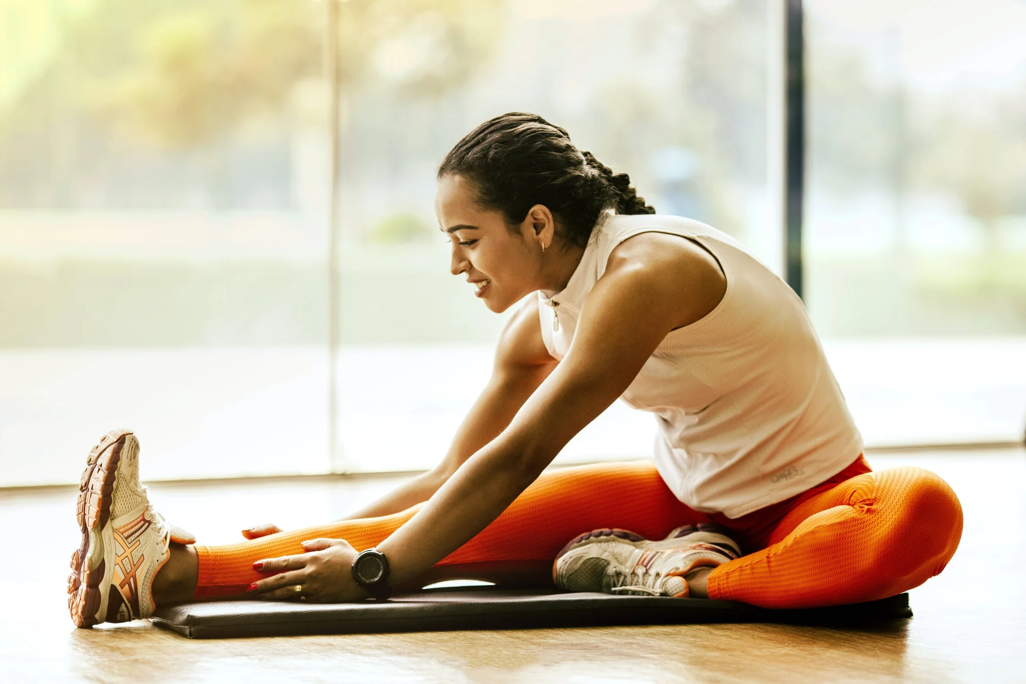 Mujer con ropa deportiva naranja realizando estiramiento de isquiotibiales sentada en una colchoneta, sonriendo.