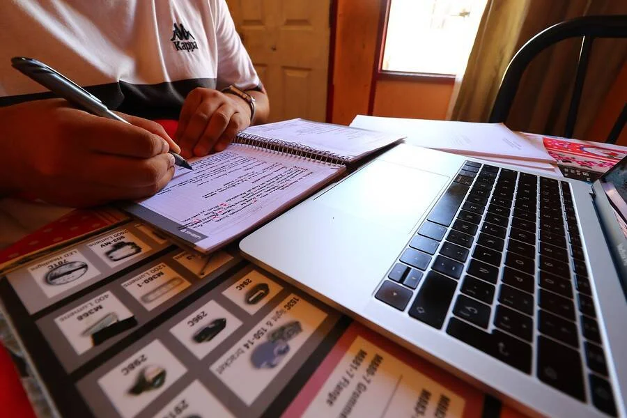 Persona escribe en cuaderno sobre una mesa con laptop, papeles y catálogos de productos.