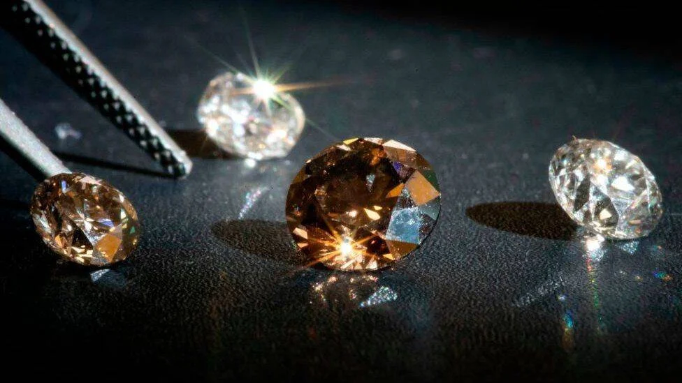 Cuatro diamantes tallados, uno de color marrón y tres incoloros, dispuestos sobre una superficie negra junto a unas pinzas.