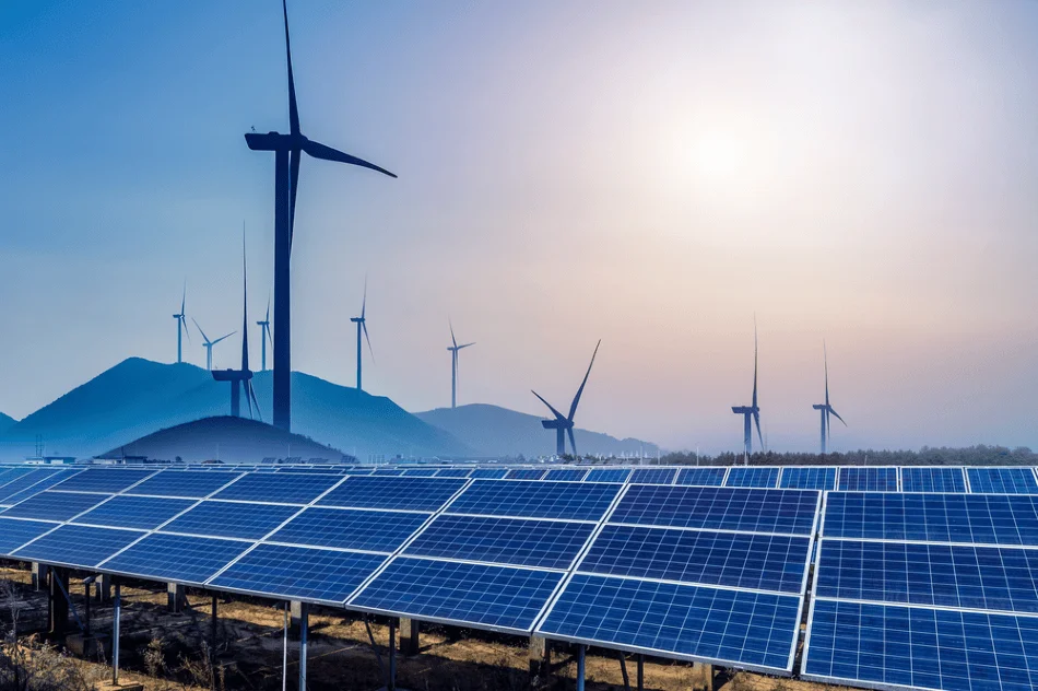 Granja de paneles solares en primer plano con aerogeneradores y montañas al fondo bajo un cielo azul.