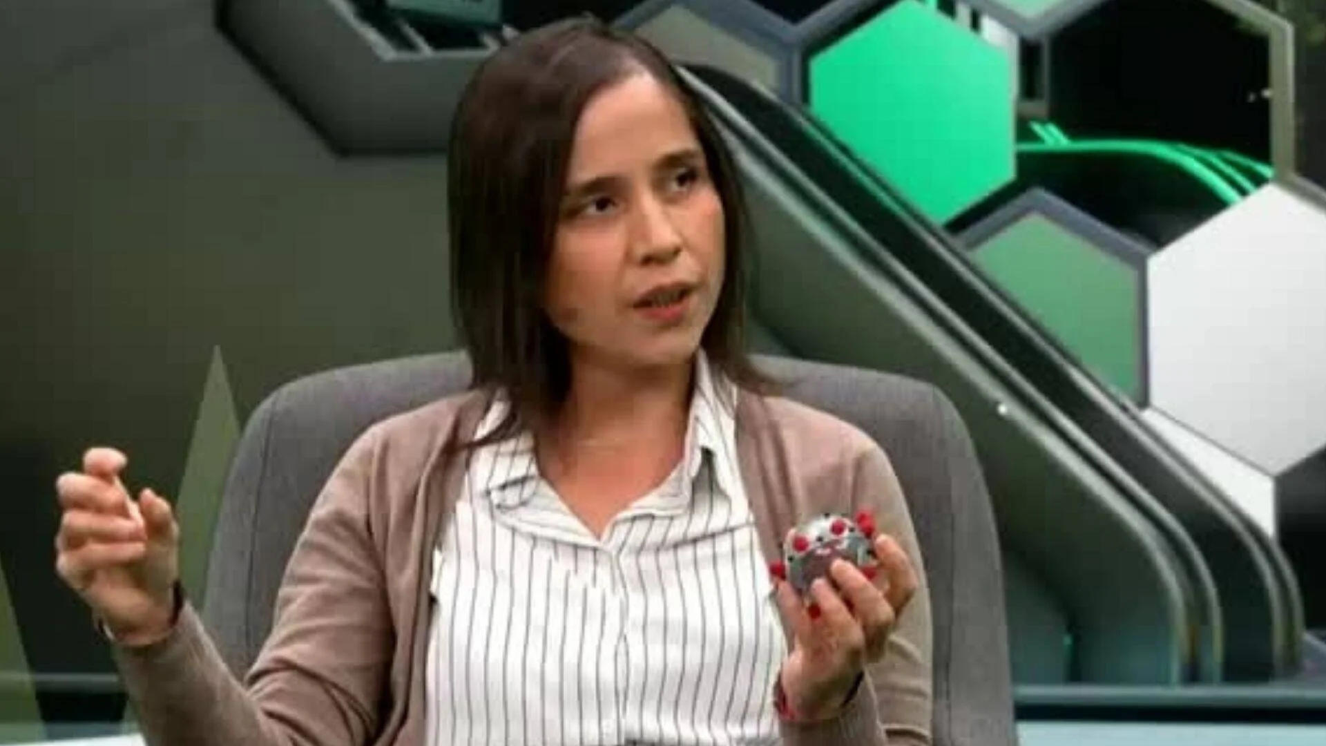 Mujer habla en un estudio de TV mientras sostiene una maqueta de partícula viral y gesticula con la mano.