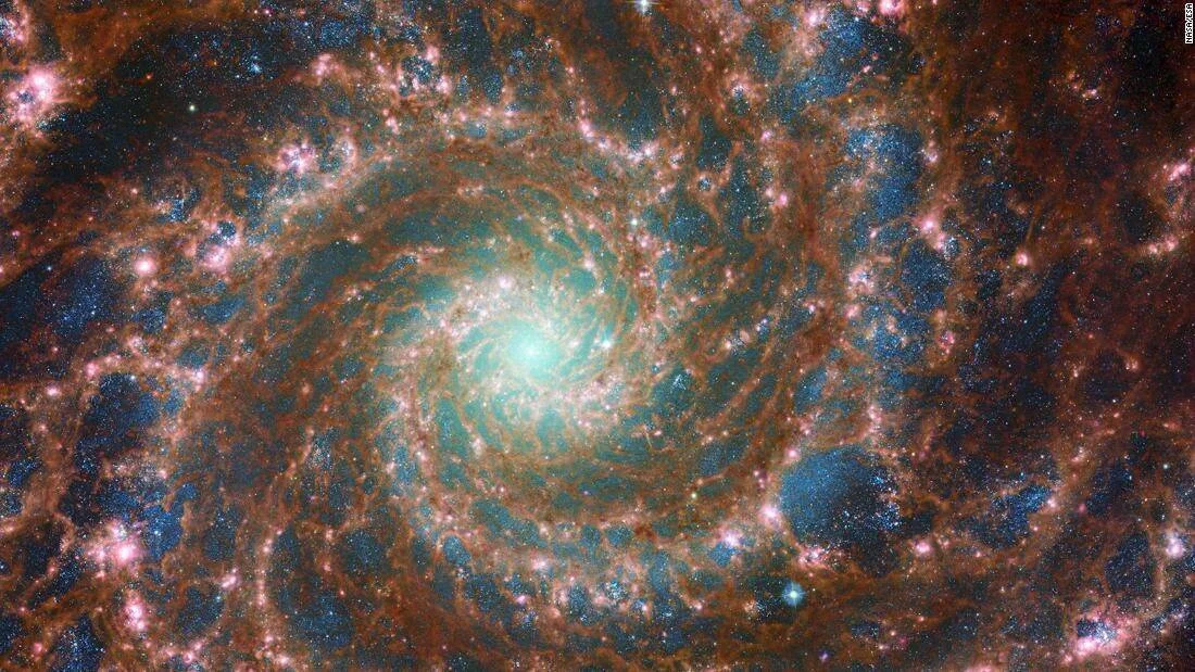 Galaxia espiral vista desde el telescopio James Webb, con núcleo turquesa y brazos de polvo marrón rojizo salpicados de estrellas.