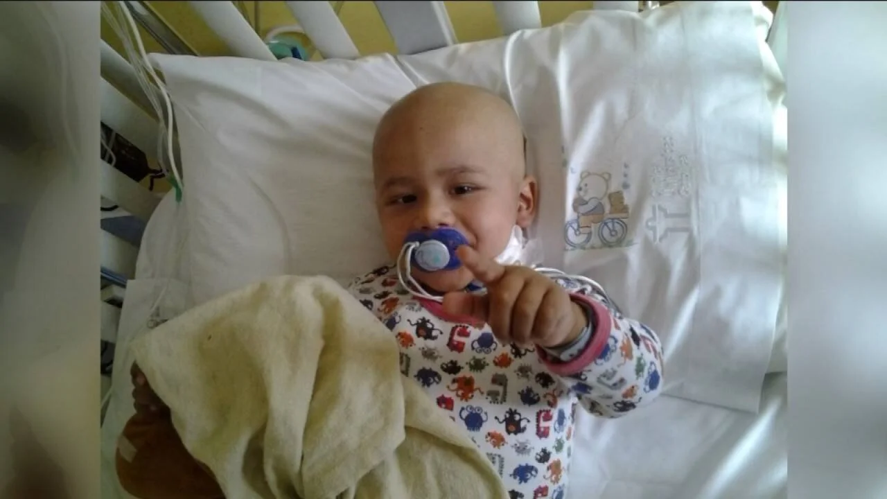 Niño pequeño sin cabello, posiblemente en tratamiento oncológico, recostado en cama de hospital con chupete en la mano.