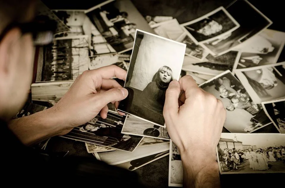 Una persona sostiene una fotografía antigua en blanco y negro de una niña, rodeada de numerosas fotos vintage dispersas sobre una mesa.
