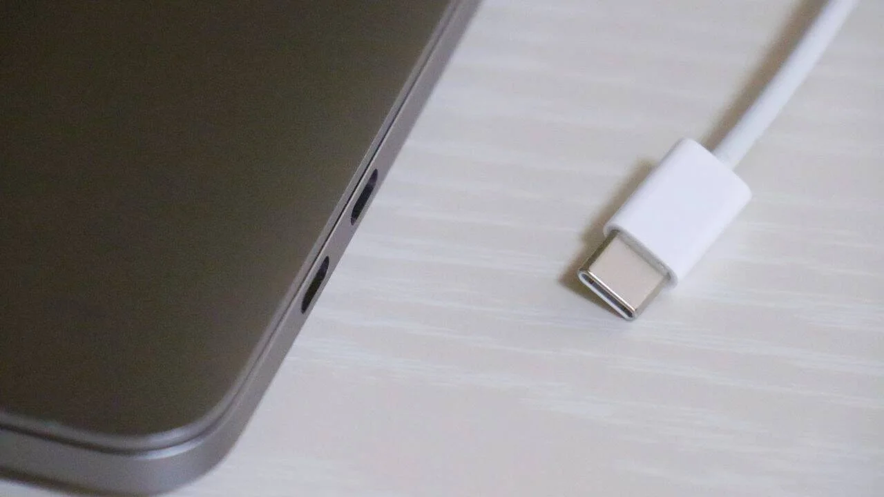 Conector USB-C blanco desconectado junto a los puertos laterales de una laptop gris sobre superficie clara.