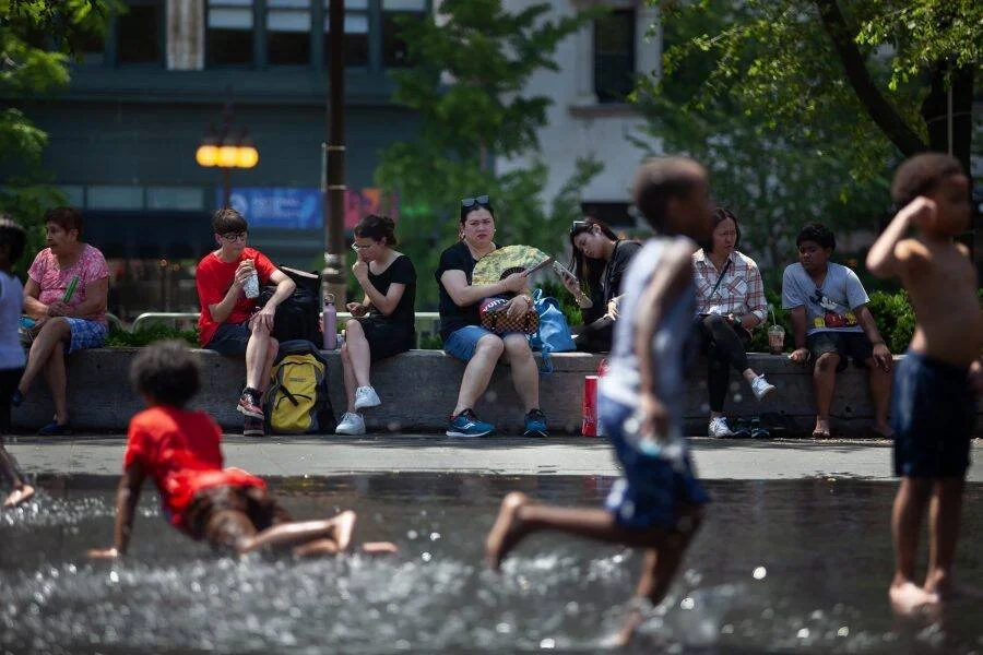 Adultos descansan en un borde de concreto mientras niños juegan en una fuente pública en un parque urbano en día de calor extremo.