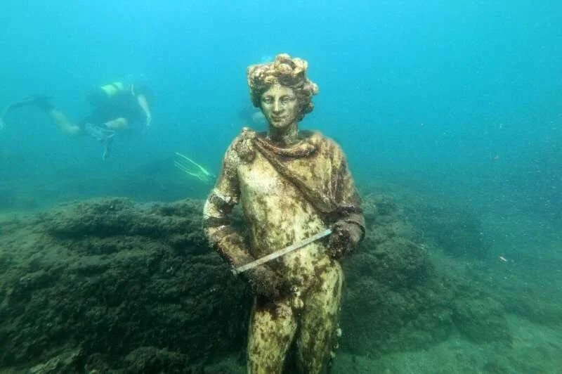 Estatua romana sumergida cubierta de sedimentos en el fondo marino, con un buzo en segundo plano.