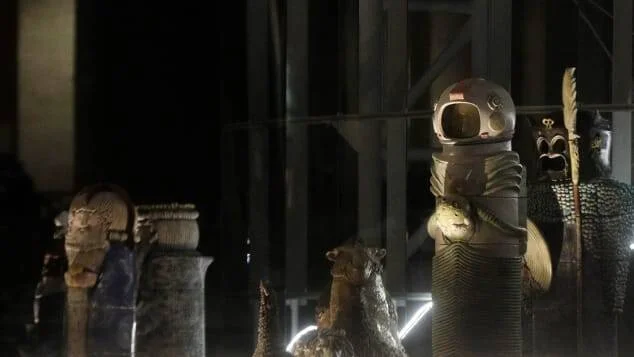 Conjunto de esculturas en penumbra que mezclan figuras precolombinas o tribales con una figura central con casco de astronauta.