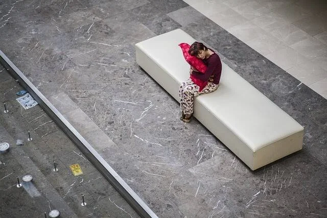 Vista cenital de una persona sentada sola en un banco blanco, abrazando una almohada roja en un espacio amplio y vacío.