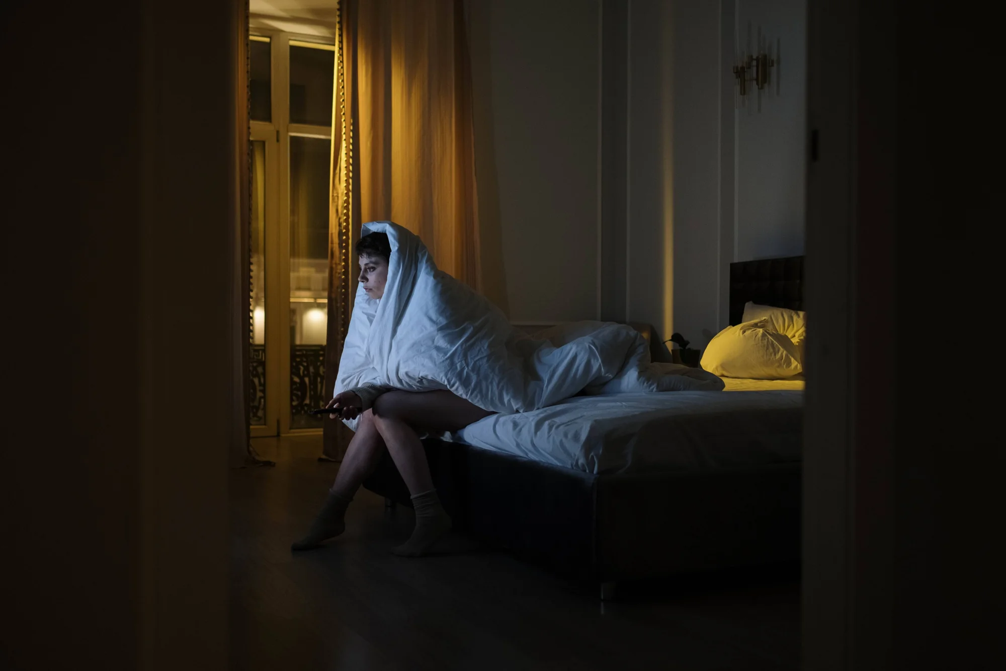 Persona sentada al borde de la cama, envuelta en una sábana blanca, mirando un dispositivo en una habitación oscura.