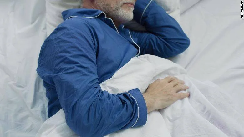 Hombre mayor con pijama azul descansando en una cama con sábanas blancas.