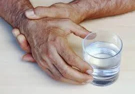 Manos de una persona mayor con temblor apoyadas sobre una mesa junto a un vaso de agua.