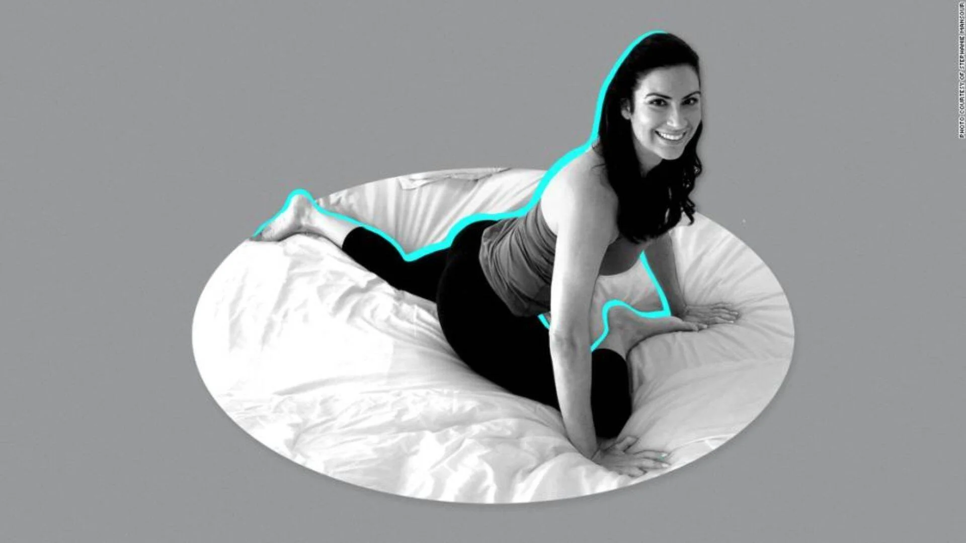 Mujer embarazada sonriente recostada sobre una almohada de maternidad en la cama, en fotografía en blanco y negro con contorno cyan.