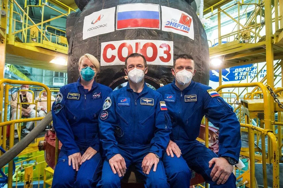 Tres astronautas con trajes azules y mascarillas posan frente a la cápsula Soyuz de Roscosmos en el hangar de lanzamiento.