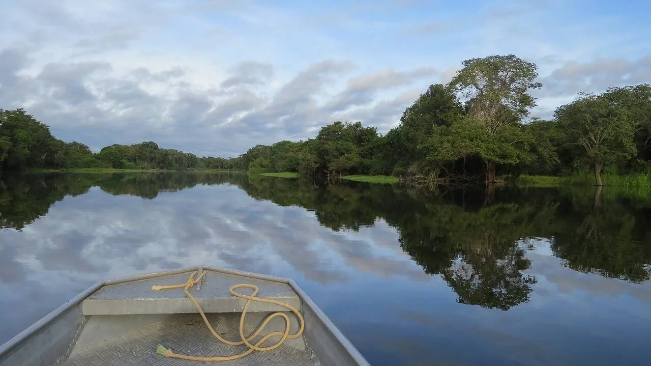 Vista desde la proa de una lancha metálica navegando un río tranquilo rodeado de selva tropical, con nubes reflejadas en el agua.